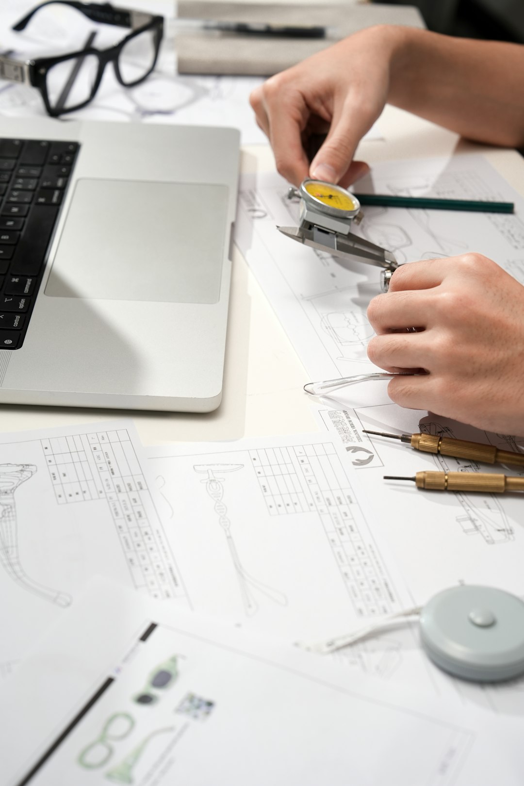 services-03 Close up of an eyewear designer using a dial caliper to measure a clear Vooglam frame on top of technical drawings beside a laptop and precision tools.